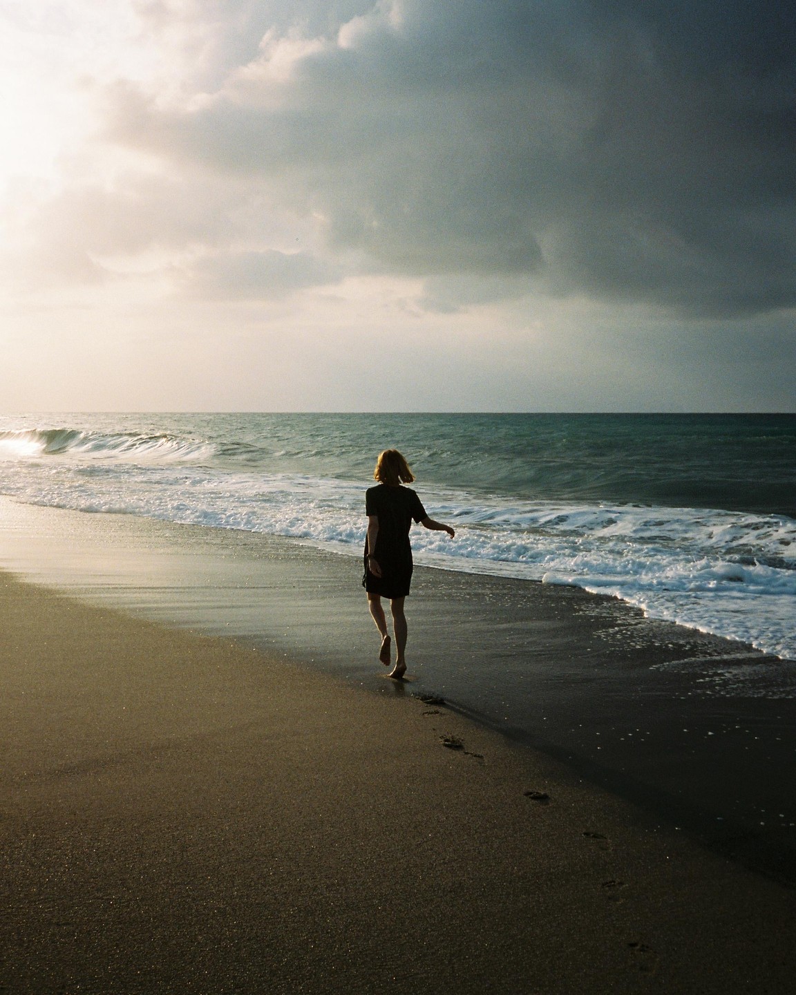 A woman walks barefoot along a beach with waves crashing on the shore under a cloudy sky.