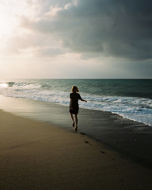 A woman walks barefoot along a beach with waves crashing on the shore under a cloudy sky.
