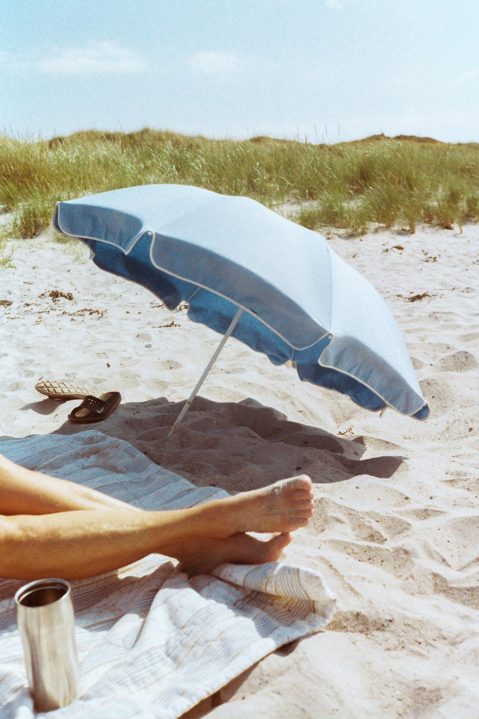 A person's legs are visible on a striped towel next to a blue beach umbrella on a sandy beach with tall grass in the background.