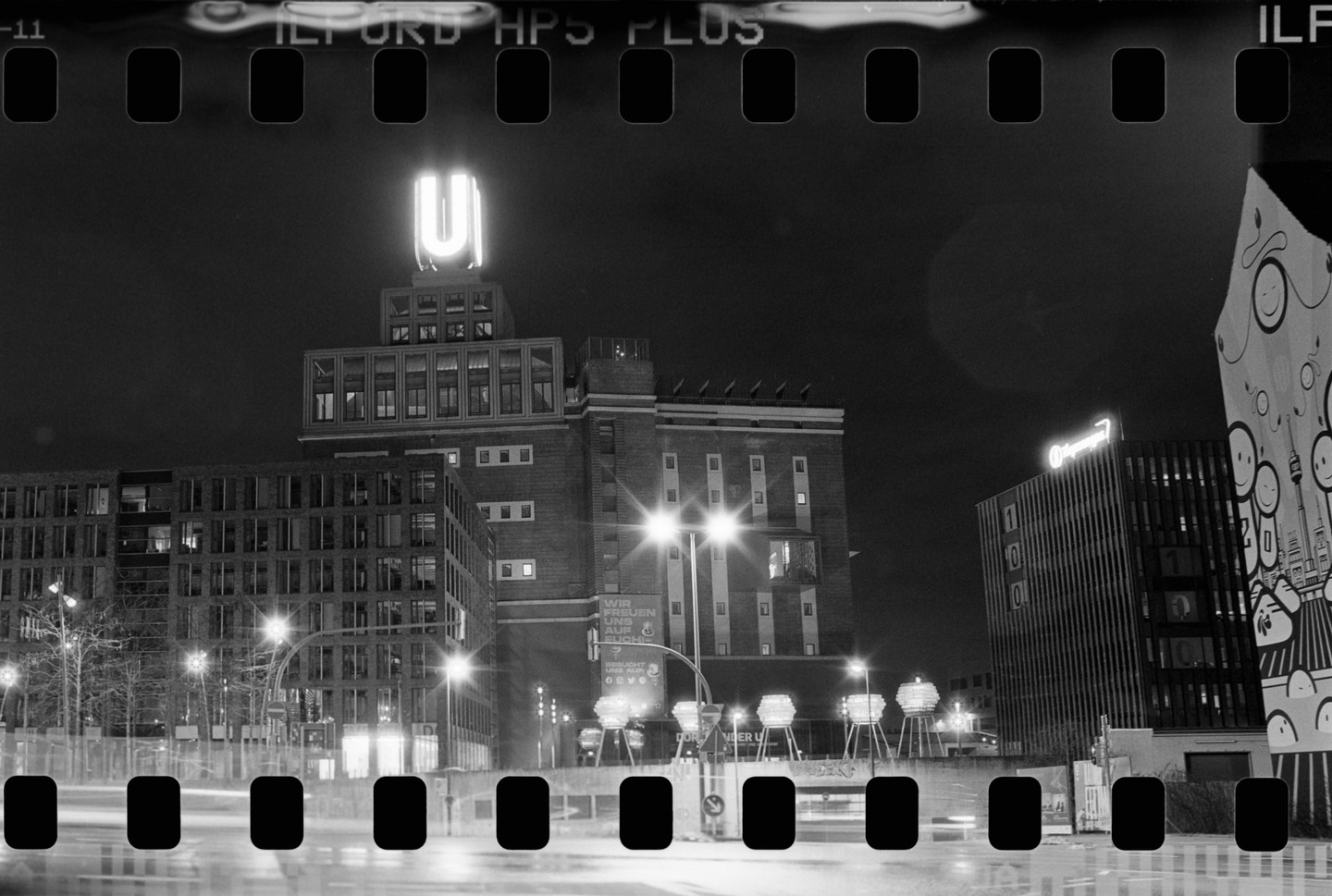 A black and white night cityscape shows a tall building with a glowing U-shaped sign on top, surrounded by other buildings and streetlights.
