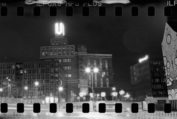 A black and white night cityscape shows a tall building with a glowing U-shaped sign on top, surrounded by other buildings and streetlights.