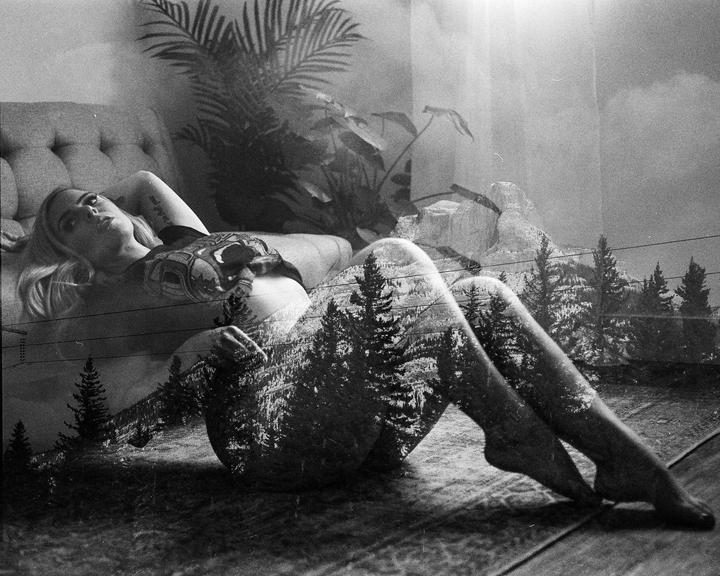 A woman reclines on a bed with a double exposure of a forest and mountains superimposed over her body.