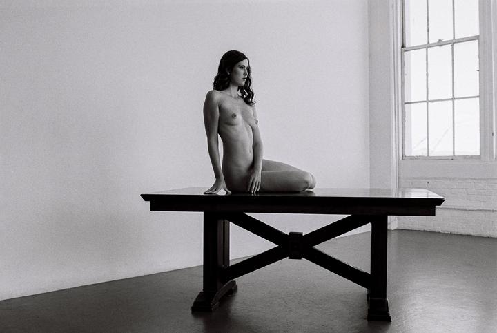 A nude woman sits on a large table in a room with a window.