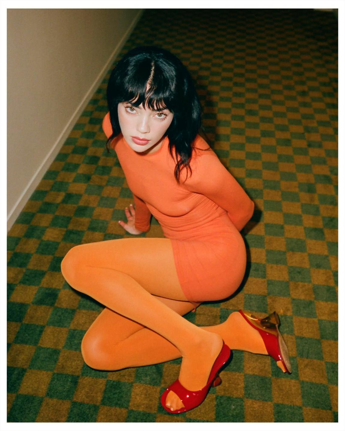 A woman in an orange dress and tights sits on a green and brown checkered carpet.