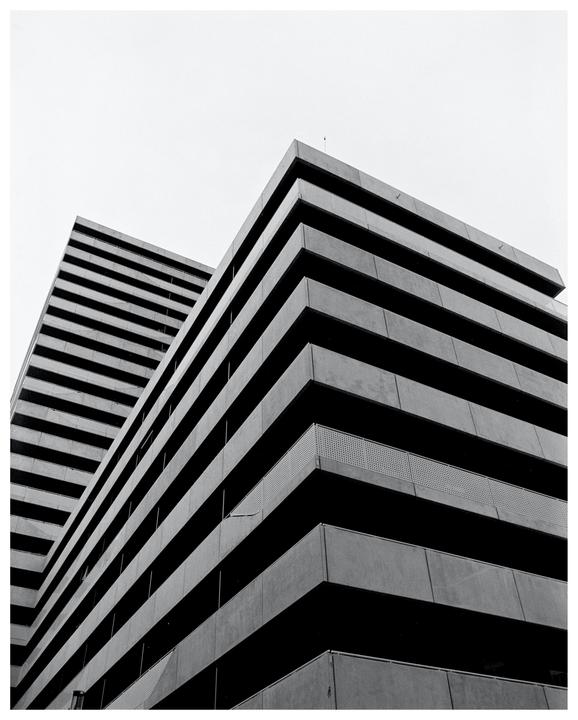 Two brutalist buildings with horizontal lines are seen from a low angle against a bright sky.