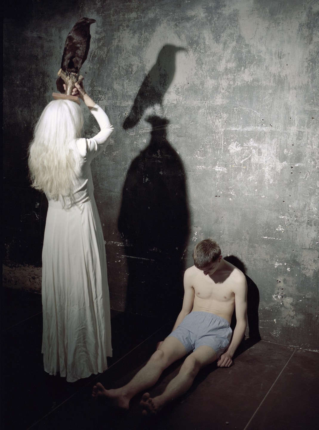 A woman in a white dress holds a taxidermy crow while a man sits on the floor next to her, with their shadows cast on a textured wall.