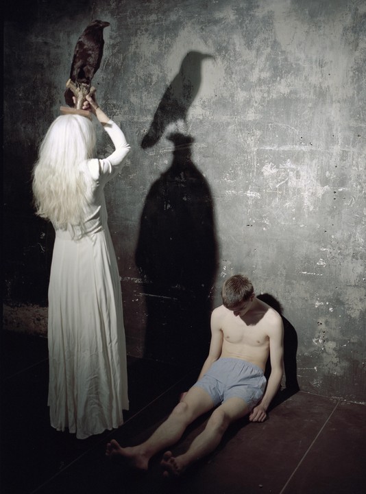 A woman in a white dress holds a taxidermy crow while a man sits on the floor next to her, with their shadows cast on a textured wall.