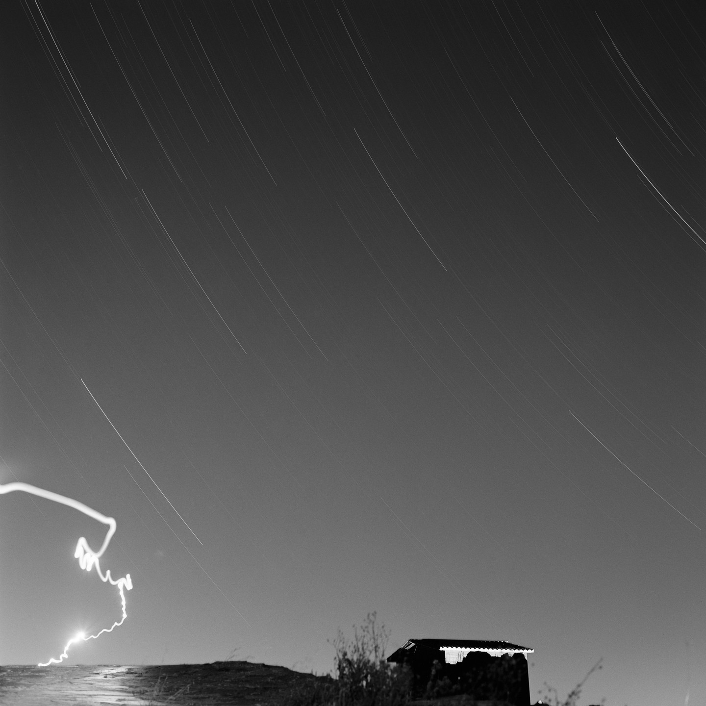 A long exposure black and white photograph shows star trails in the night sky above a small structure on a hill.