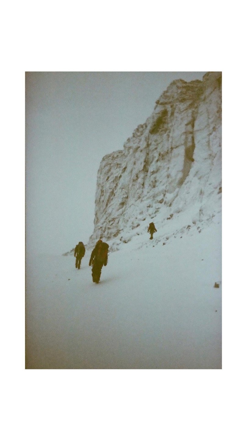 Three people walk through a snowy landscape with a large rock formation on the right.