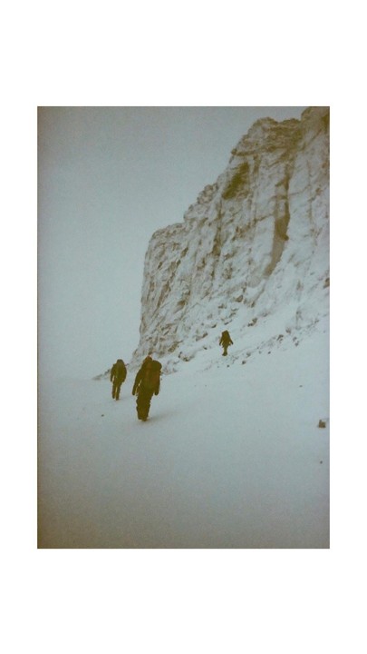Three people walk through a snowy landscape with a large rock formation on the right.