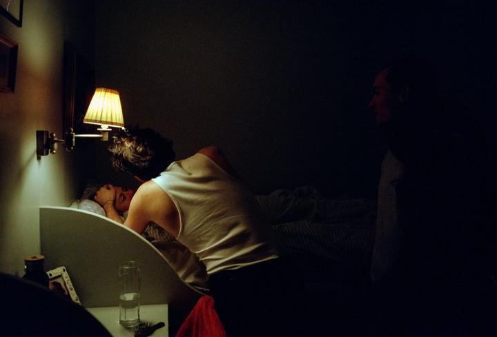 A man leans over a woman in bed, illuminated by a bedside lamp, while another man sits in shadow.