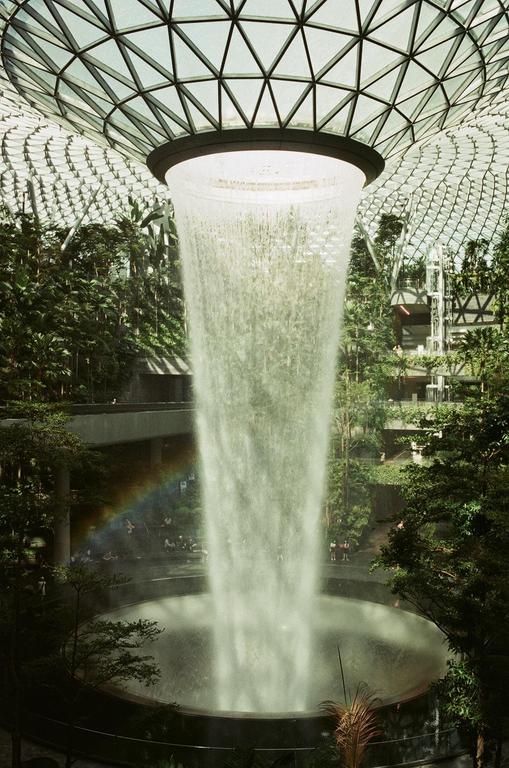 A large indoor waterfall cascades from a circular opening in a glass-domed ceiling into a pool below, surrounded by lush greenery and a visible rainbow.