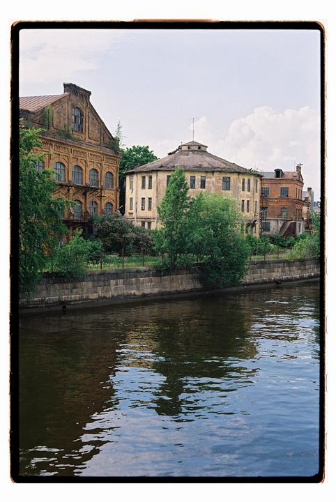 A river flows past old buildings and green trees under a cloudy sky.