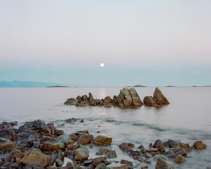 The moon is visible above a body of water with several rocky formations in the foreground and distant land in the background.