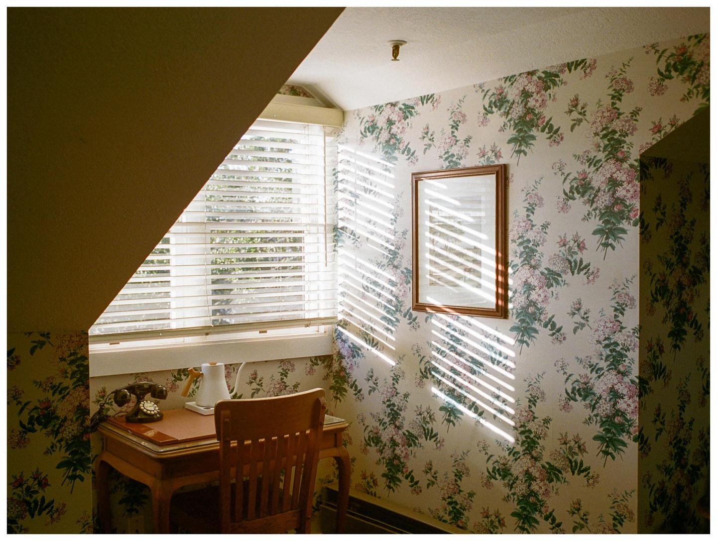 Sunlight streams through window blinds onto a floral wallpapered wall with a framed picture and a wooden desk with a telephone and kettle.
