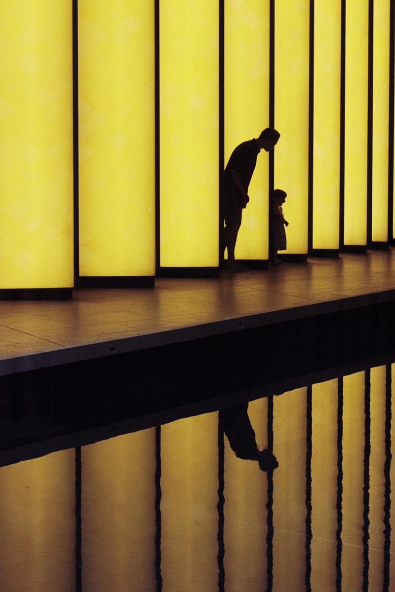 A man and a child are silhouetted against a wall of yellow light panels, with their reflections visible in a dark surface below.