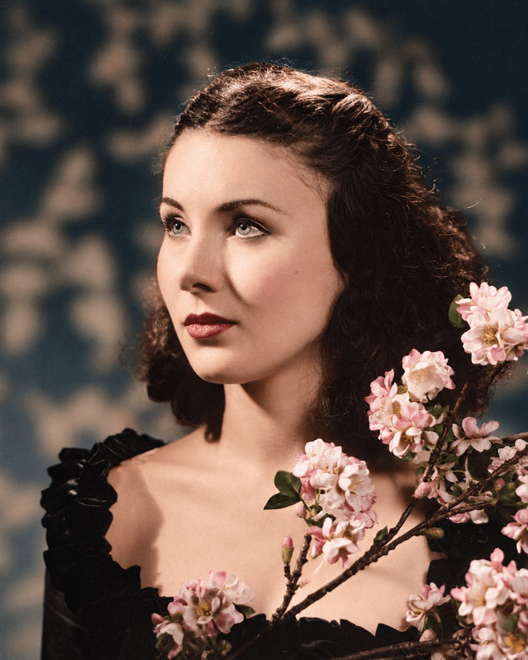 A woman with dark hair and red lips is looking up and to the left, with pink blossoms in the foreground.