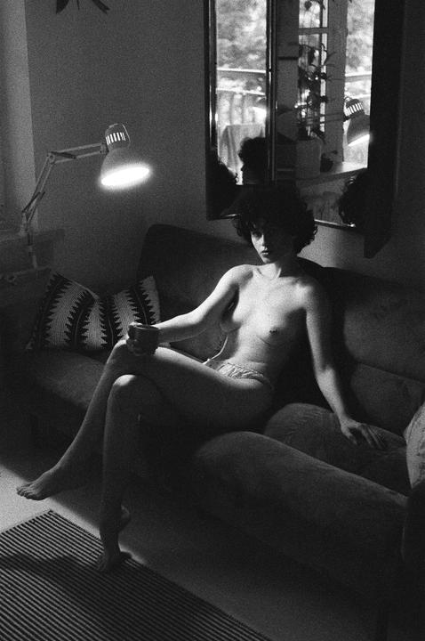 A woman with curly hair sits on a sofa, holding a cup, with a lamp and a mirror visible in the background.
