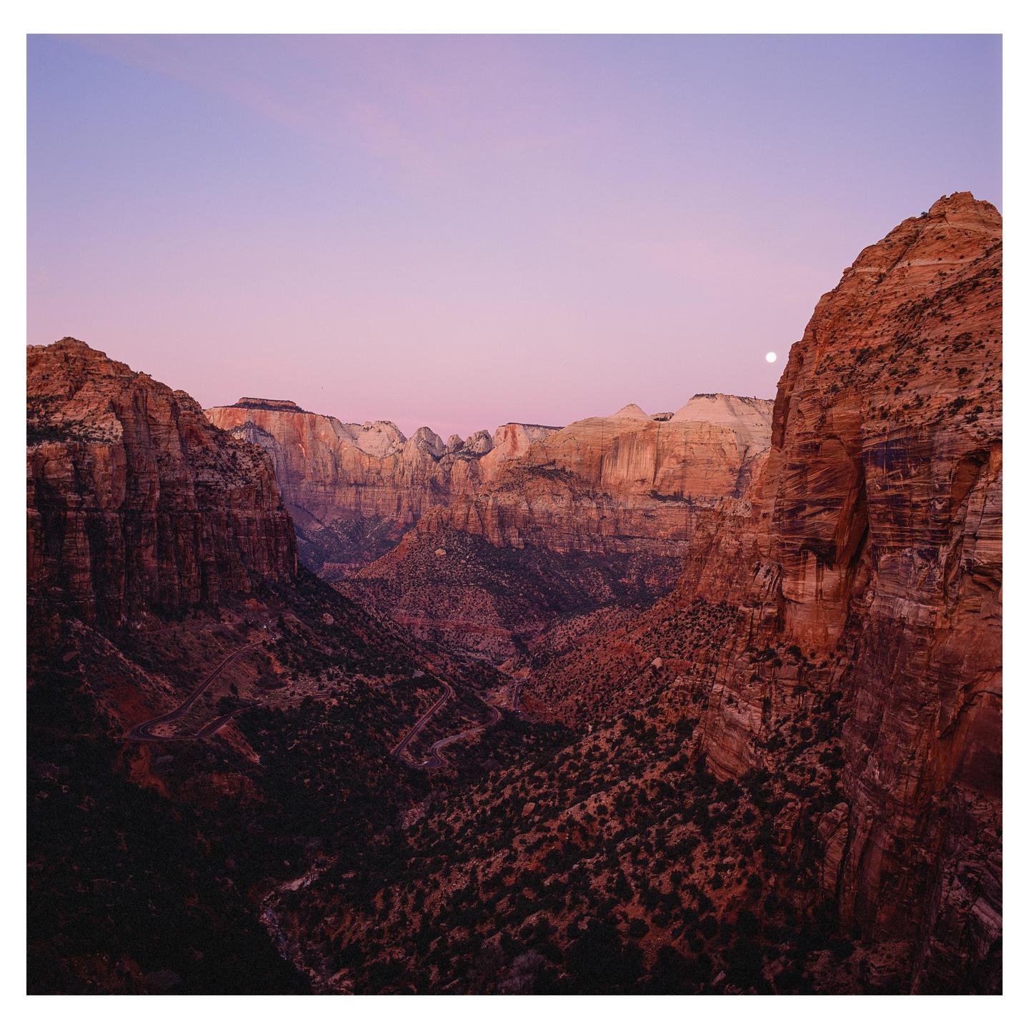 A full moon is visible over a canyon landscape with a winding road at dusk.