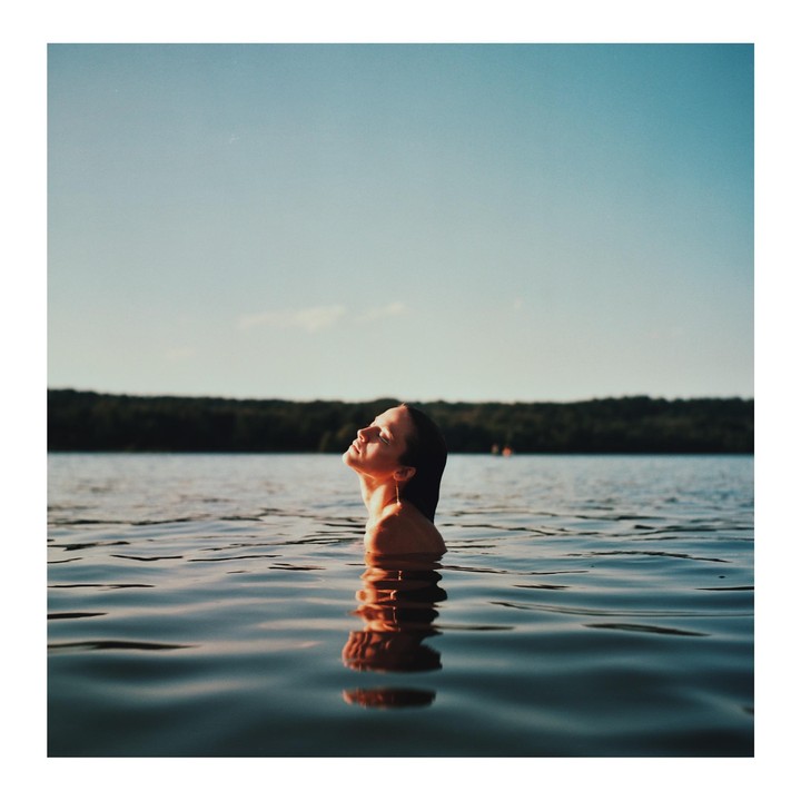 A woman with her head tilted back is in a body of water with a forest in the background.