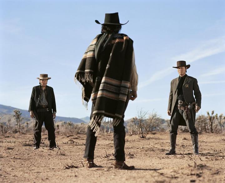 Three men in cowboy hats and western attire stand in a desert landscape under a clear sky.