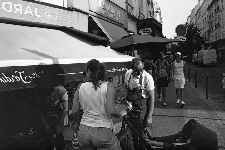 A man in an apron stands next to a woman pushing a stroller on a city street with storefronts and other people.