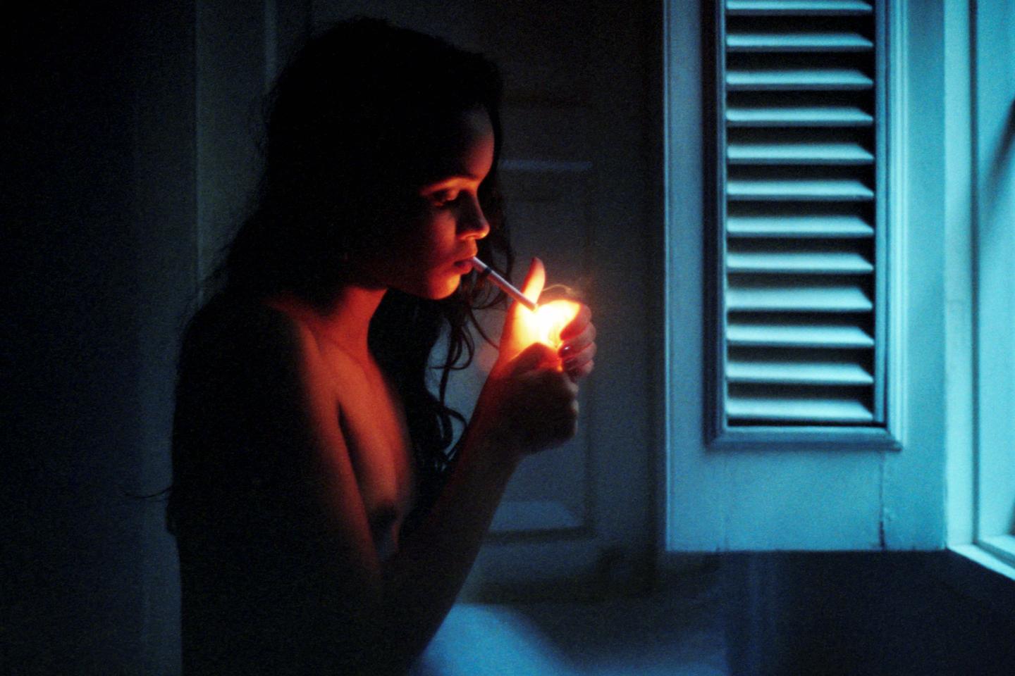 A woman lights a cigarette in a dimly lit room with a window to her right.