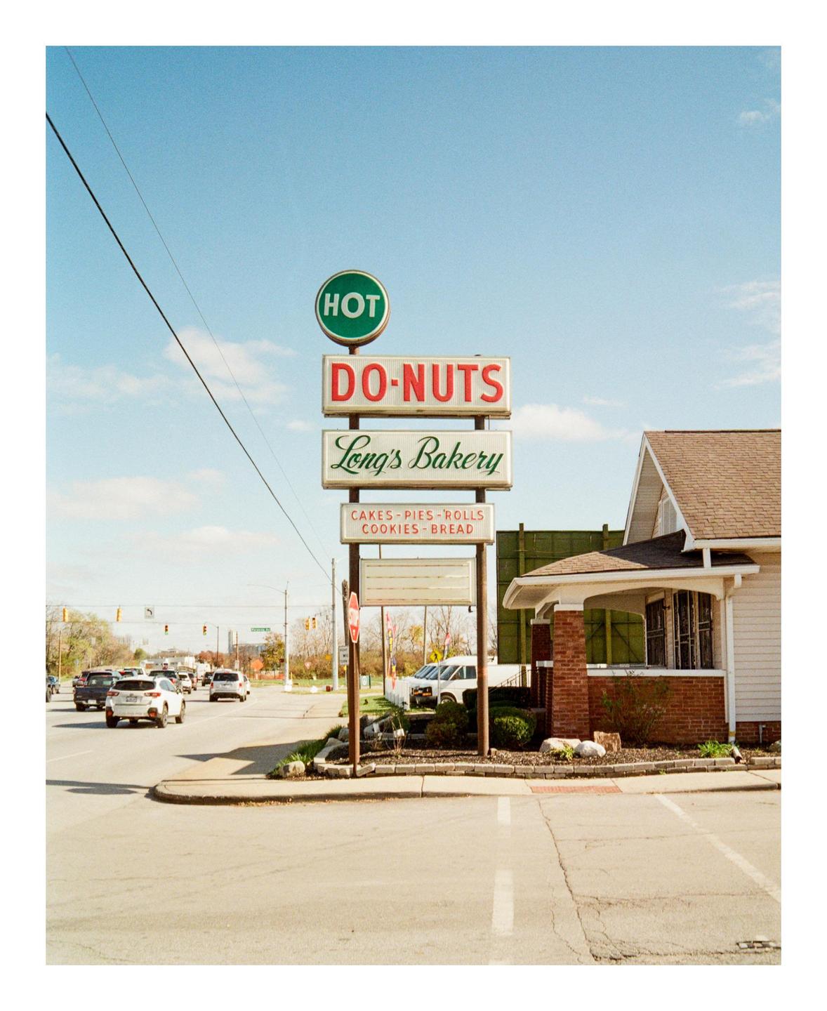 A sign for "Hot Do-Nuts Long's Bakery" stands next to a building at a street intersection with cars.