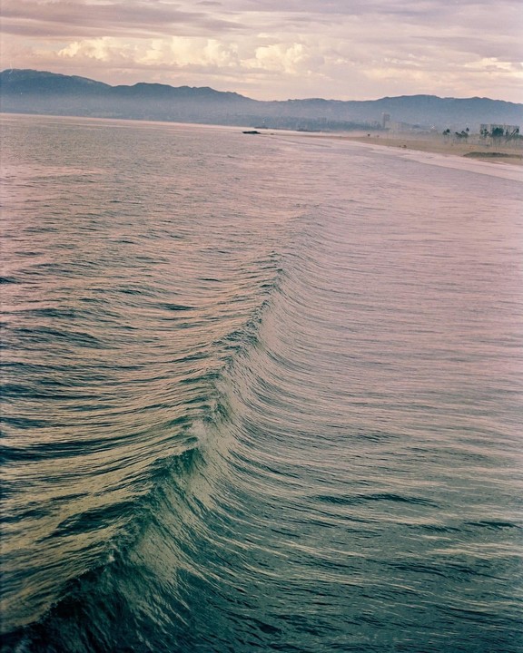 A single wave rolls towards a distant beach with mountains and a hazy sky in the background.