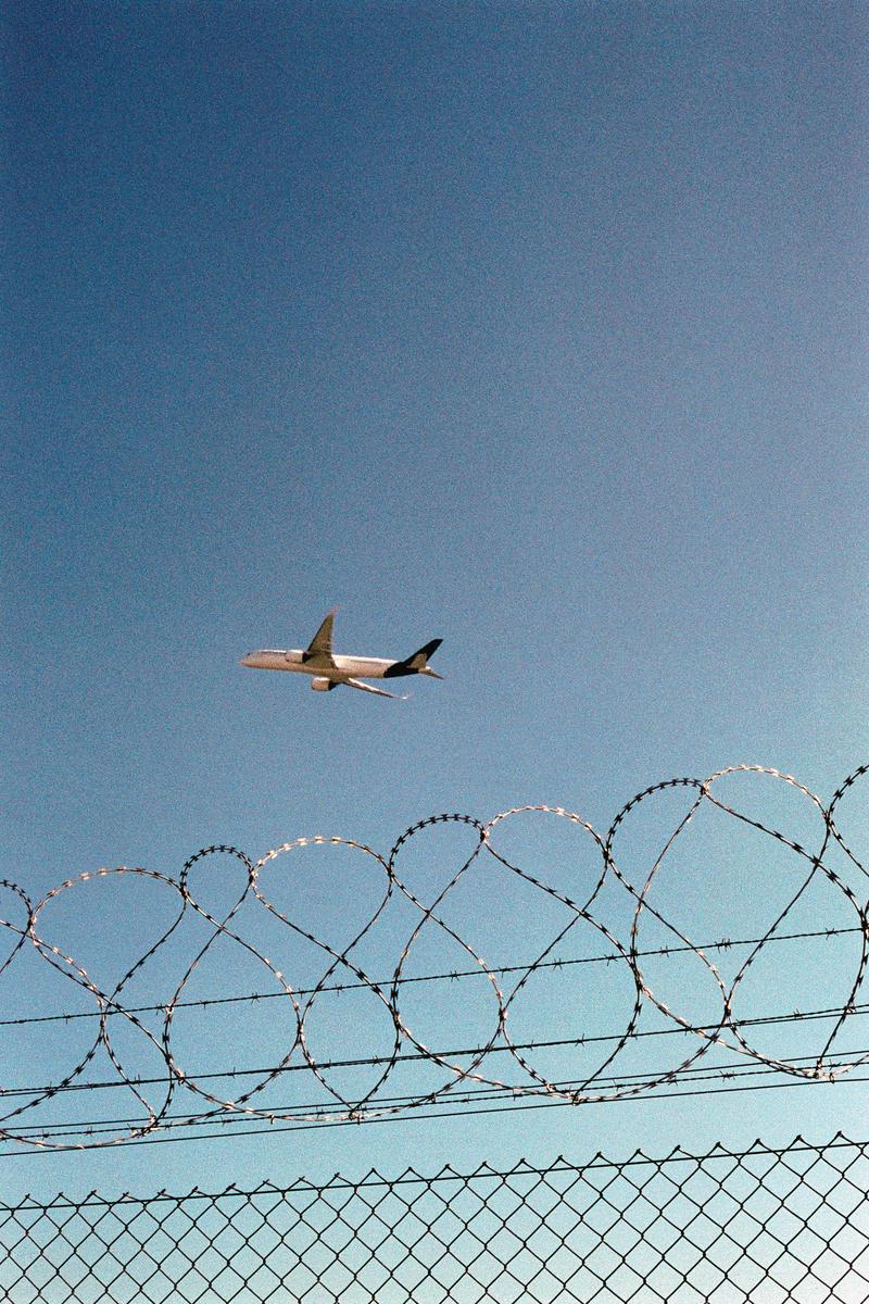 A passenger airplane flies in a clear blue sky above a chain-link fence topped with razor wire.