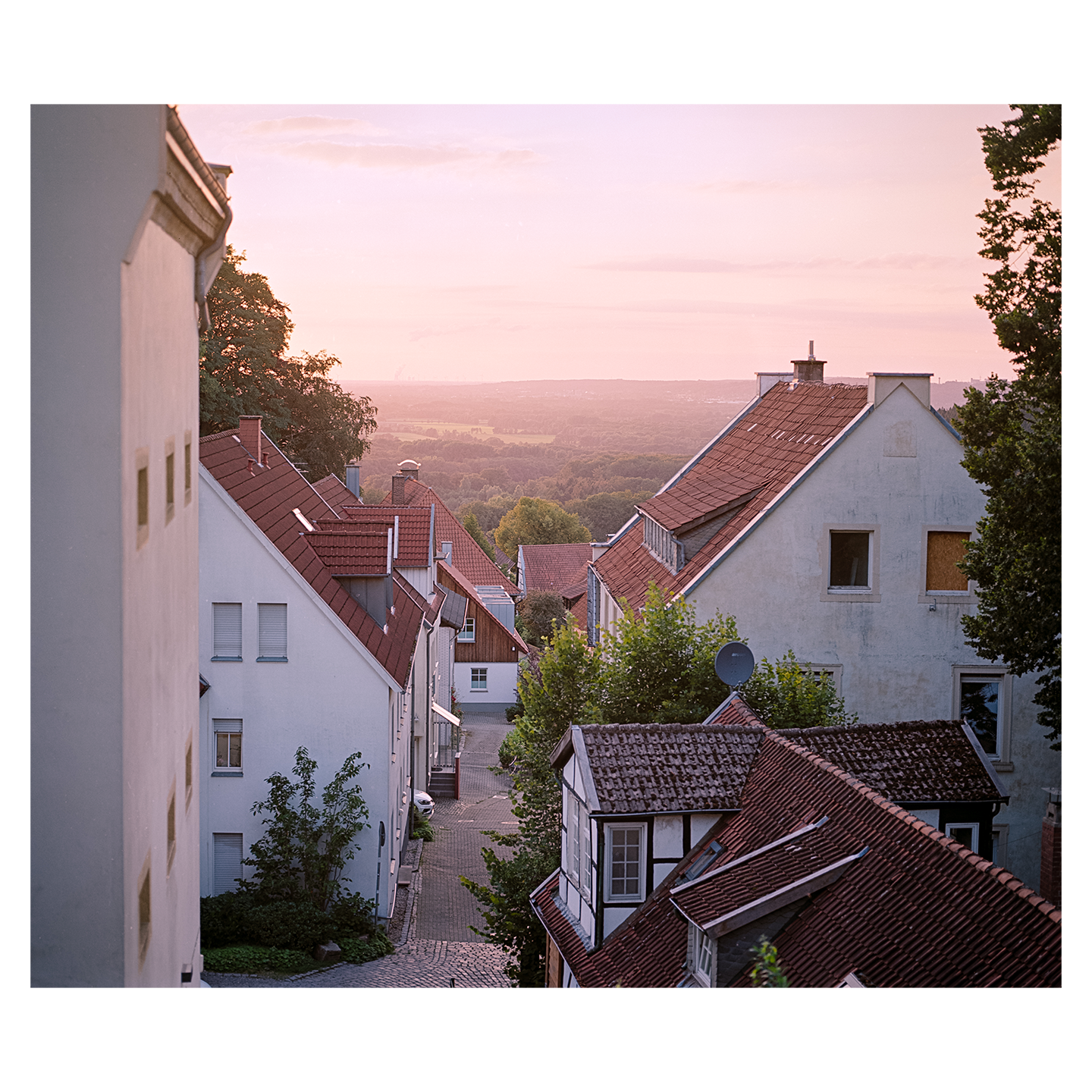 A view of a village with red-tiled roofs and white buildings, looking down a street towards a valley at sunset.