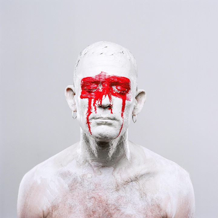 A man with white paint covering his head and torso has red paint streaked across his closed eyes and dripping down his face.