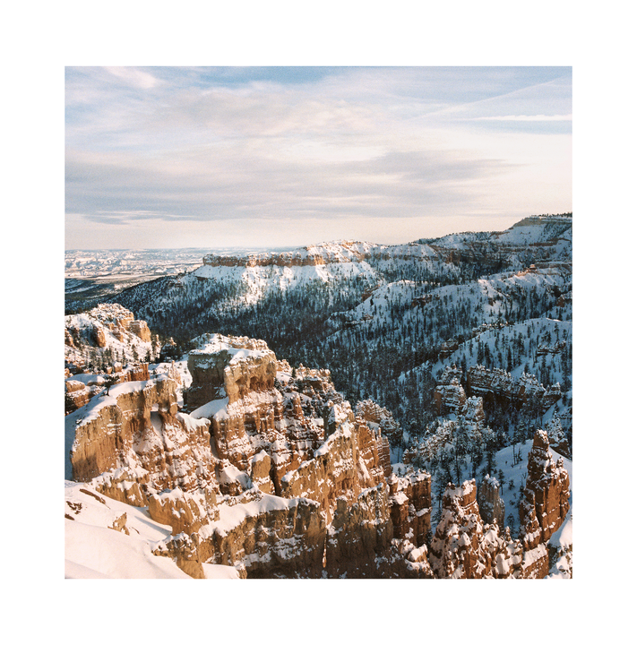 A snowy landscape of Bryce Canyon shows red rock formations and pine trees under a cloudy sky.