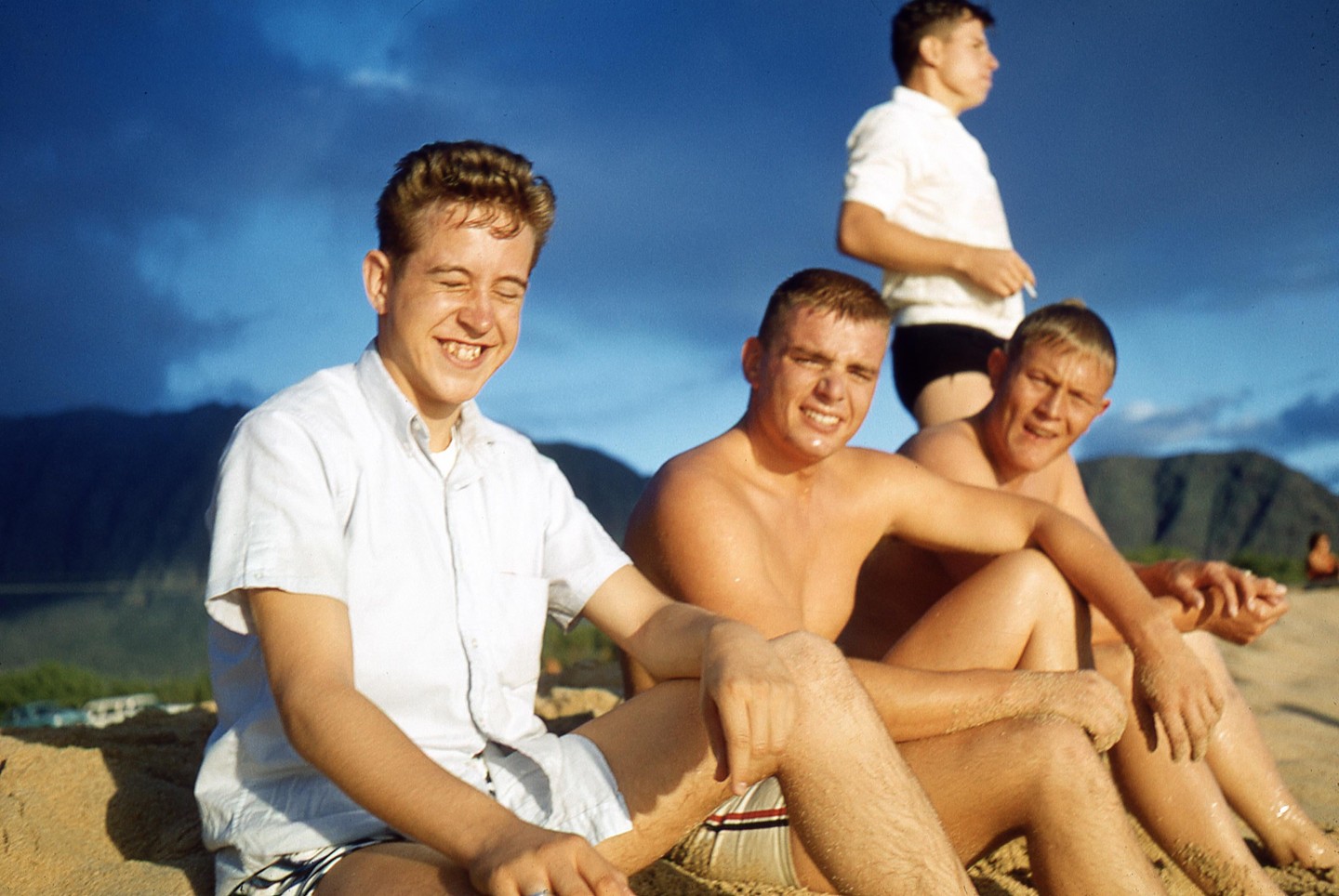 Four young men are sitting on a sandy beach with mountains in the background under a blue sky.