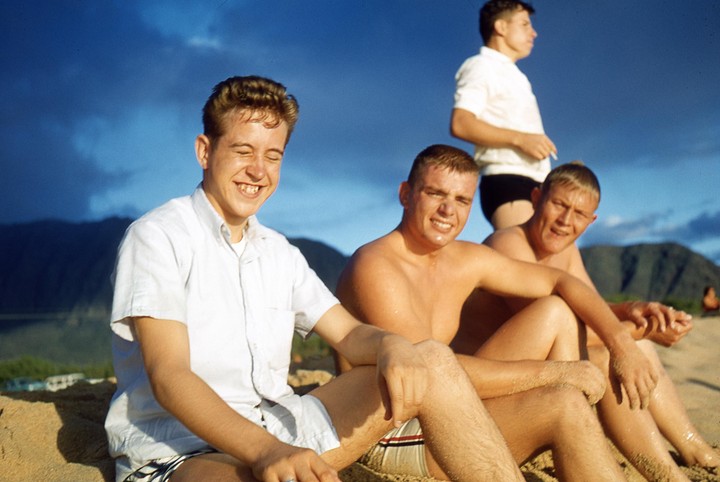 Four young men are sitting on a sandy beach with mountains in the background under a blue sky.