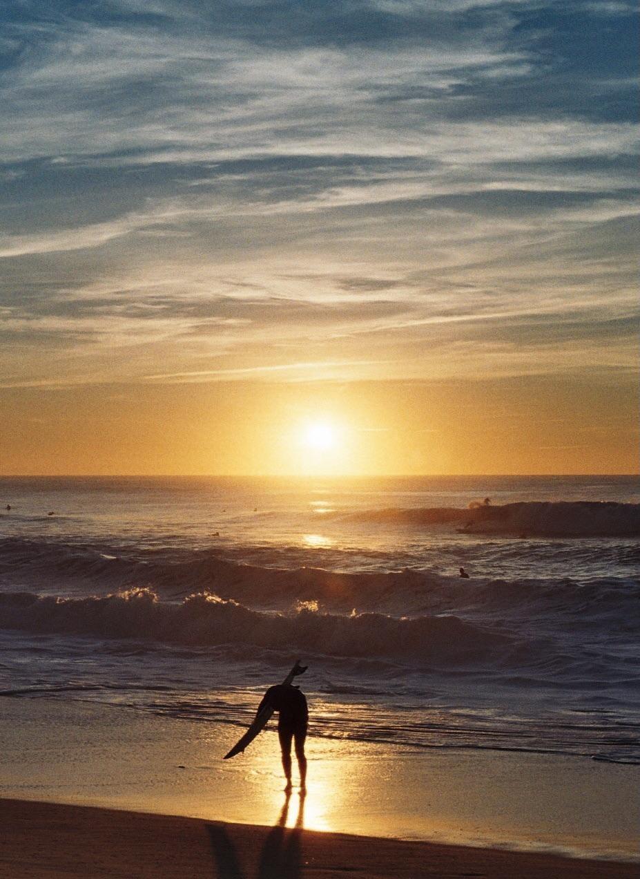 A surfer stands on the beach at sunset, holding a surfboard and looking out at the ocean waves.