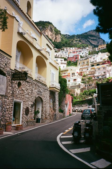 A narrow street winds past a stone building with a hotel sign, leading up to a hillside town with many colorful houses.
