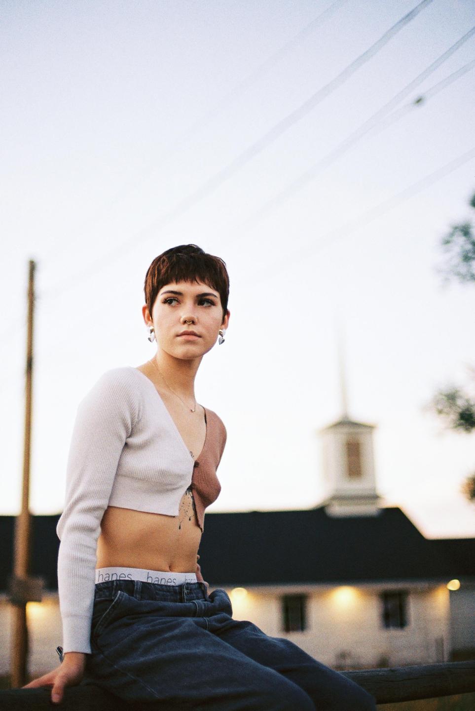A woman with short hair and a crop top sits outdoors with a building and power lines in the background.