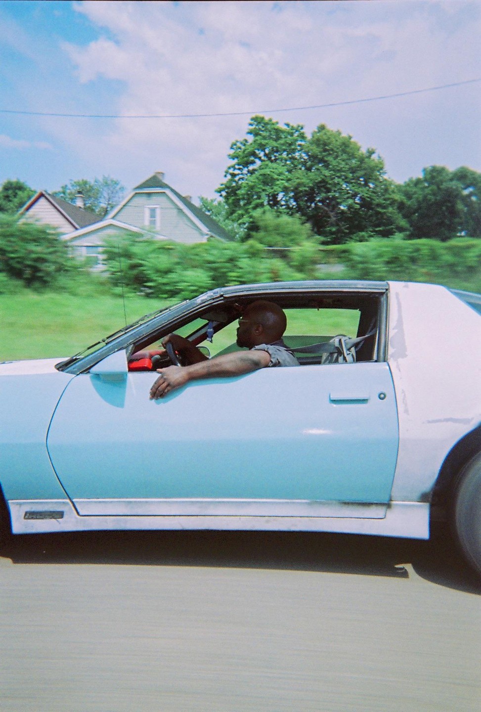 A man drives a light blue car past houses and trees.