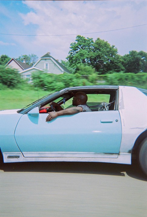 A man drives a light blue car past houses and trees.