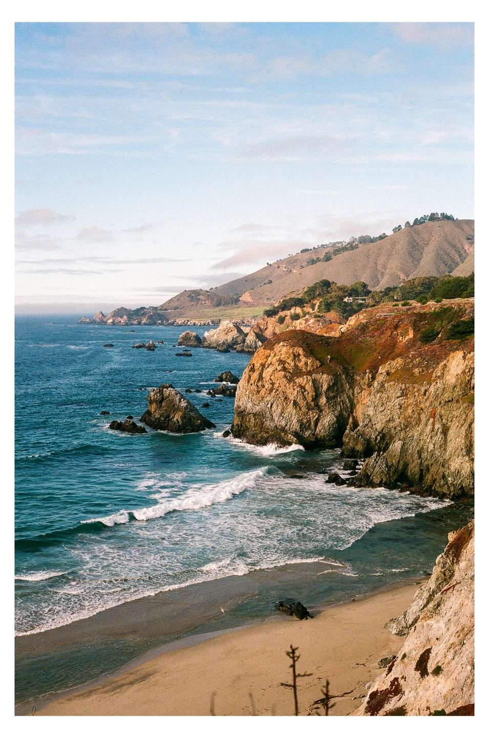 Waves crash on a sandy beach next to a rocky cliff with a mountain in the background.
