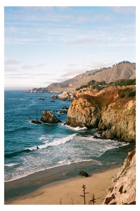 Waves crash on a sandy beach next to a rocky cliff with a mountain in the background.
