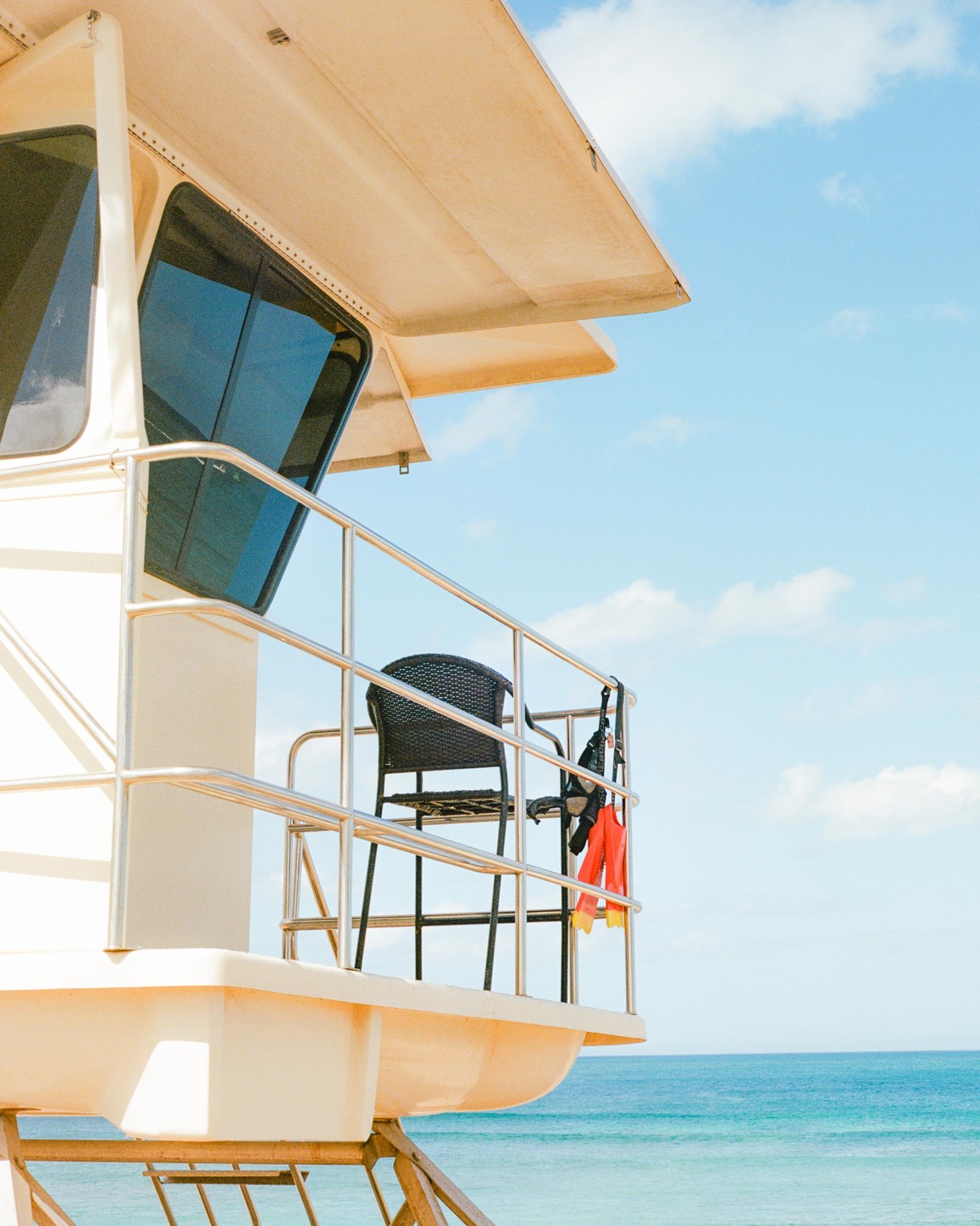 A lifeguard stand with a chair and fins hanging from the railing overlooks a calm blue ocean under a partly cloudy sky.