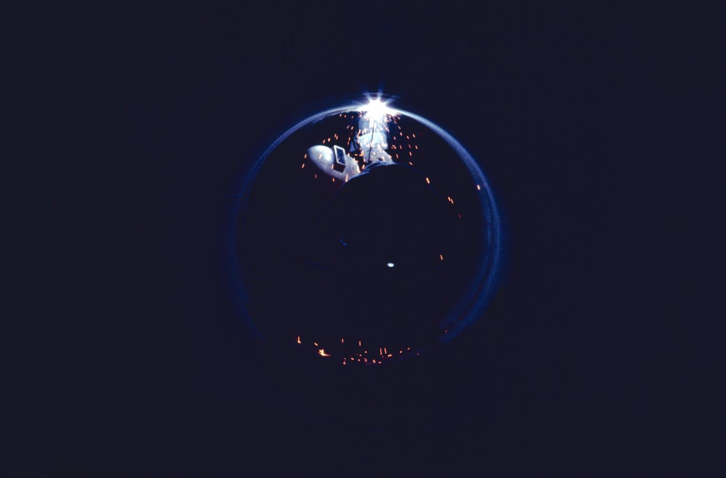 A welder works on a pipeline at night, surrounded by sparks and a circular light trail.