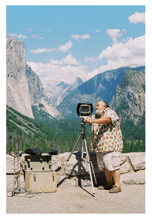 A man with a beard is operating a large format camera on a tripod, overlooking a mountain valley.