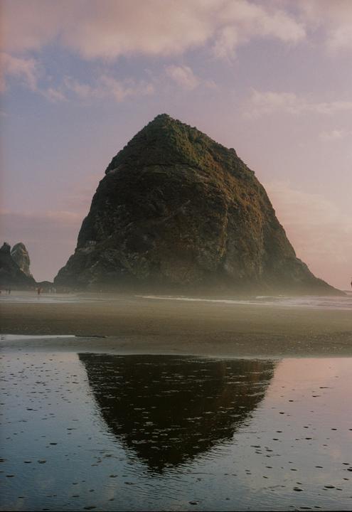 A large rock formation stands on a beach with its reflection visible in the wet sand.