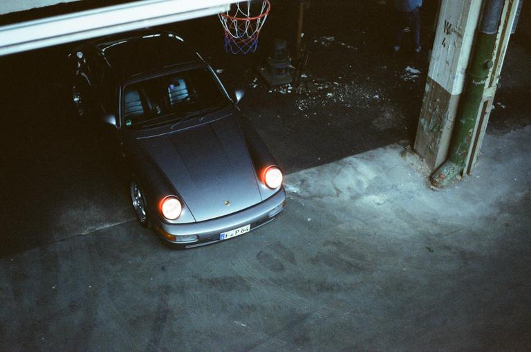A gray car with its headlights on is parked in a garage with a basketball hoop visible above it.