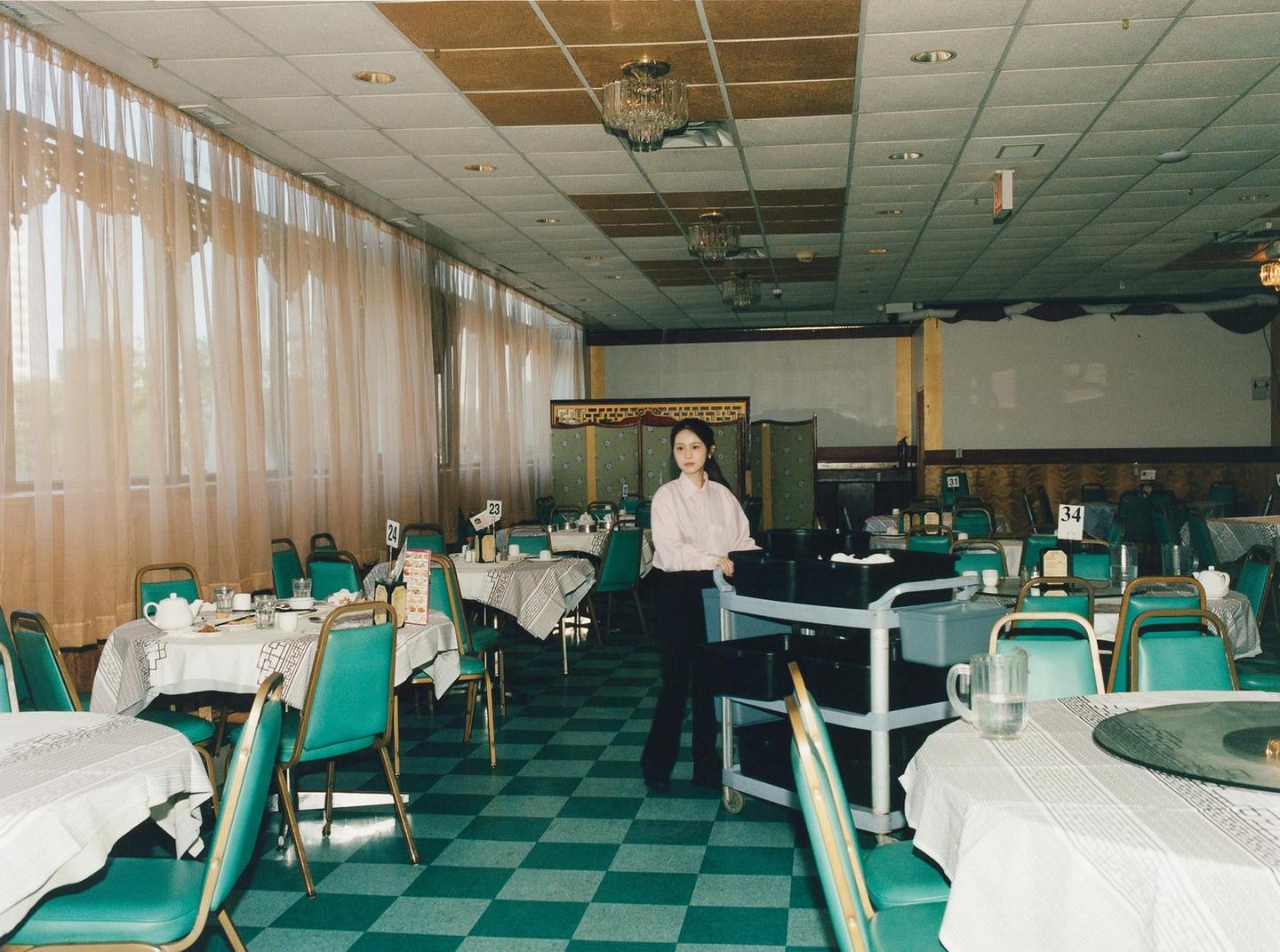 A woman stands next to a serving cart in a large, empty dining room with many tables and chairs.