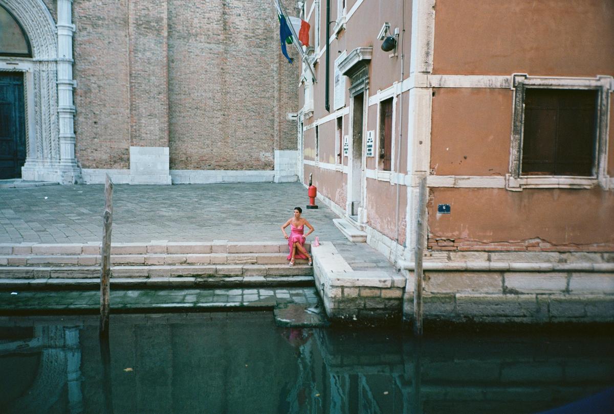 A woman in a pink dress sits on the edge of a canal in Venice.