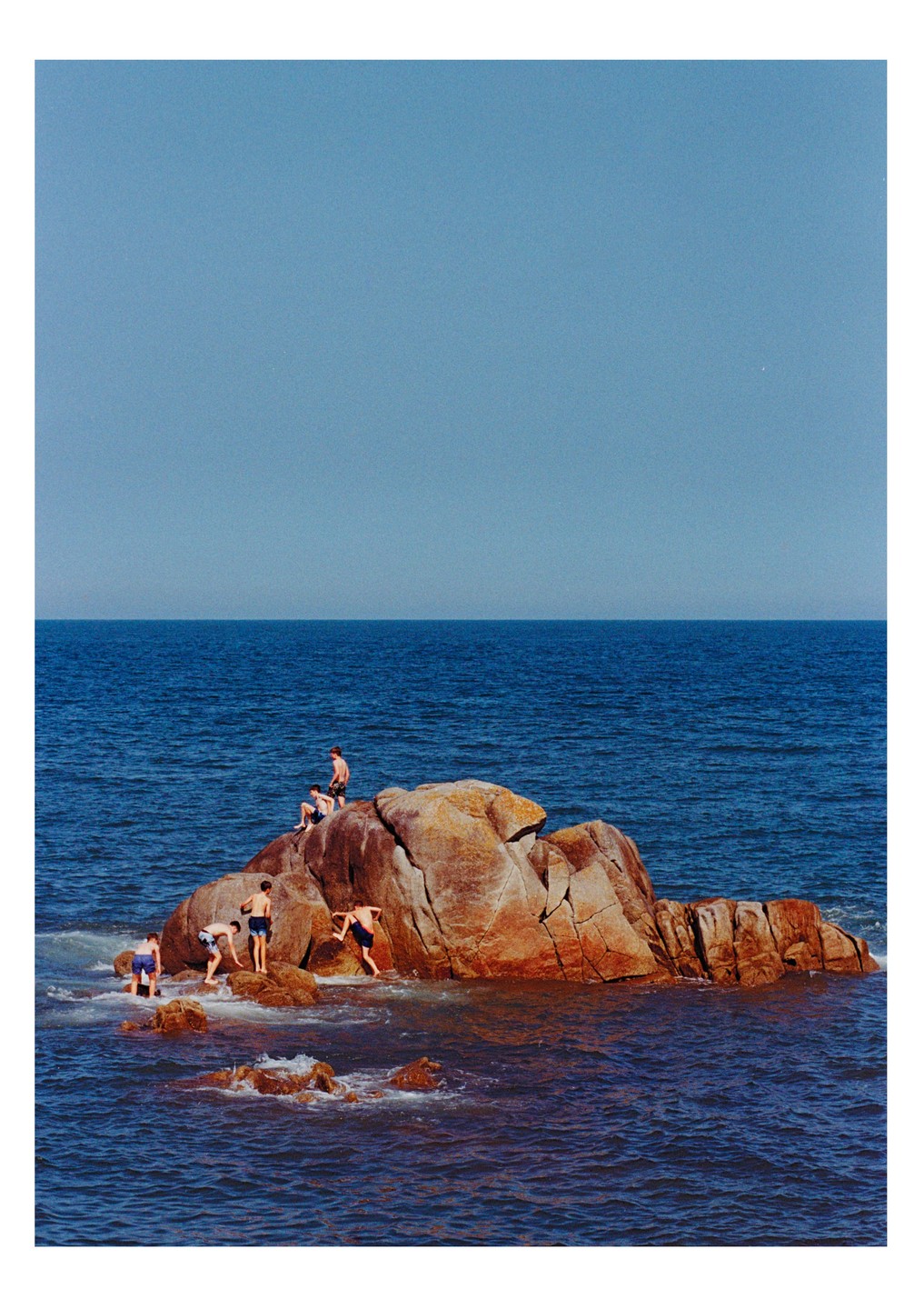 Several boys are playing on and around a large rock formation in the ocean under a clear sky.